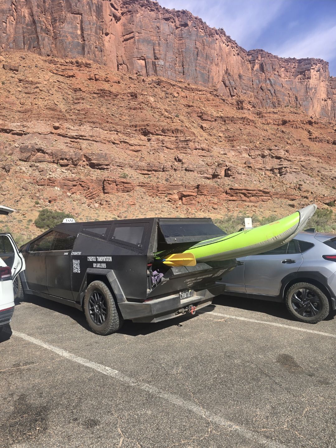 The CyberTaxi Cybertruck with a kayak loaded in the bed, parked beneath Moab red rock cliffs